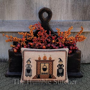 May include: A rectangular cross-stitch sampler on beige fabric featuring two figures in pointed hats, a brick fireplace with a hanging black cauldron, three orange pumpkins on the mantel, and two black cats, all framed by a thin black border.