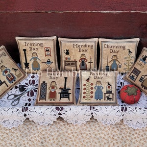 May include: Nine small embroidered pillow covers with a vintage aesthetic. Each pillow cover features a different scene depicting a woman performing a daily chore, such as washing, ironing, mending, churning, baking, resting, and cleaning. The pillow covers are displayed on a wooden tray with a lace doily and a red tomato pincushion.