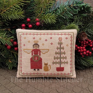 May include: A cross-stitched Christmas pillow featuring an angel, a cat, and a Christmas tree. The angel wears a red dress and has a halo. The pillow is beige with red and green accents. The text "The Humble Stitcher" is at the bottom.