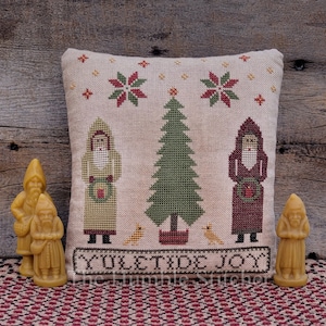 May include: A cross-stitch pillow with a Christmas tree, two figures in red and yellow robes, and the text "Yuletide Joy". The pillow is on a wooden surface with four small, yellow, gnome-like figures.