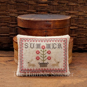 Pode incluir: Uma almofada retangular bordada em ponto cruz com a palavra "SUMMER" bordada na parte superior com linha preta. Abaixo da palavra, há flores vermelhas em um vaso preto. A almofada está em uma caixa de madeira. The Humble Stitcher é impresso na parte inferior.