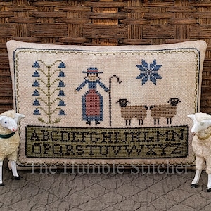 May include: A beige pillow with a cross-stitch design of a woman with a shepherd's crook, three sheep, a flower, and a tree. The pillow has the alphabet embroidered on the bottom and the text "The Humble Stitcher".