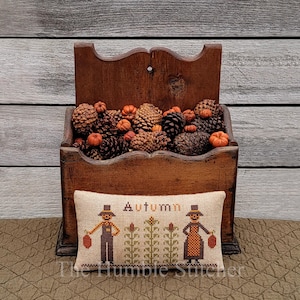 Op de afbeelding: Een houten wandplank gevuld met dennenappels en kleine oranje pompoenen. Een rechthoekig kussen met het woord "Autumn" en kruissteekfiguren van een man en een vrouw die pompoenen vasthouden, staat voor de plank.