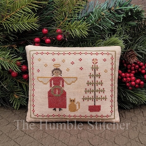 Pode incluir: Um bordado em ponto cruz quadrado sobre tecido bege claro com um anjo de túnica vermelha e asas douradas, um pequeno gato amarelo e uma árvore de Natal verde estilizada. O design é emoldurado por uma borda geométrica vermelha com pequenas estrelas douradas.