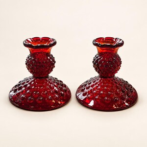 May include: A pair of matching ruby red glass candle holders featuring a textured hobnail pattern across the wide circular bases and the central stems. Each holder has a scalloped rim at the top opening, designed to securely hold a standard taper candle.