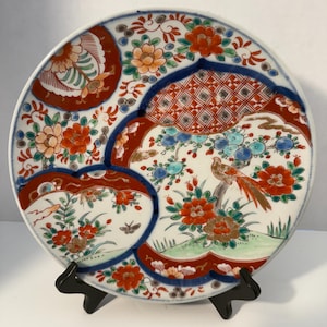 May include: A round ceramic plate featuring a detailed hand-painted design with a central bird perched among orange and green flowers. The surface is divided into sections by deep blue borders, incorporating red geometric patterns and small butterflies.