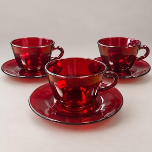 May include: Three matching translucent ruby red glass teacups and saucers arranged in a triangular formation. Each cup features a rounded bowl shape with a curved handle, and each saucer has a circular indentation to hold the cup securely.