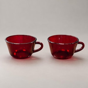 May include: Two matching translucent ruby red glass cups, each featuring a wide, rounded bowl shape and a curved side handle. The glass has a smooth, polished finish that catches the light, creating deep red tones throughout the structure.