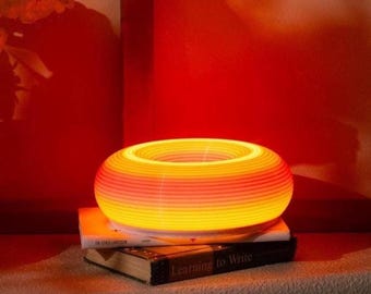 May include: A glowing, circular lamp with horizontal stripes of orange and yellow, resting on two books. The lamp emits a warm light, creating a cozy ambiance. The books are titled "On Lars Larsson" and "Learning to Write".