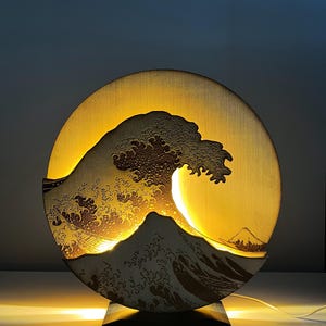 Puede incluir: Lámpara redonda de madera con un diseño detallado de "La Gran Ola de Kanagawa" de Hokusai. El diseño está cortado con láser y retroiluminado con una luz amarilla cálida, creando una silueta llamativa de la ola y el monte Fuji.