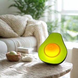 May include: A green avocado-shaped lamp with a glowing yellow center sits on a wooden table. The lamp is lit, casting a warm glow. The background includes a couch, a woven basket, and greenery.