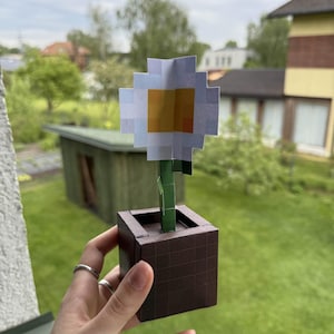 May include: A pixelated paper flower in a brown square pot. The flower has white petals and a yellow center. The green stem rises from the pot. The pot is held in front of a green background.