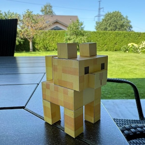 May include: A papercraft llama figurine, constructed from tan and yellow patterned cardboard. The llama has a blocky, pixelated design, with rectangular legs, body, and head. The figurine is posed on a dark, textured surface, with a green lawn and trees in the background.