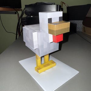 Puede incluir: Un modelo de pollo de papel con un diseño pixelado, con un cuerpo blanco y gris, un pico marrón y una cresta roja. El pollo se apoya en patas amarillas y está colocado sobre una base rectangular blanca. El modelo es de papel y está sobre una superficie gris.
