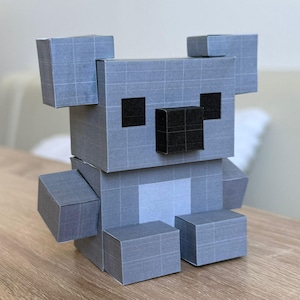 May include: A gray, geometric koala bear figure made of paper. The koala has square ears, a black nose, and black eyes. The body is constructed from multiple rectangular blocks, giving it a pixelated appearance. The koala is sitting on a wooden surface.