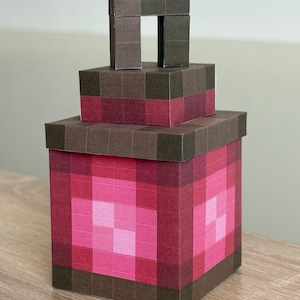 Pode incluir: Uma estrutura de caixa pixelizada de três níveis em tons de castanho e rosa. A caixa maior é quadrada, com uma tampa e uma estrutura semelhante a uma alça na parte superior. O design apresenta um padrão em blocos e pixelizado, que lembra a estética de um videogame.