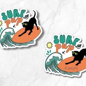 Op de afbeelding: Een sticker met een zwarte hond met een zonnebril die surft op een oranje surfboard. De sticker heeft de woorden "Surf Pup" in teal en oranje, met een zon en een golf. De naam van de hond, "Kona", staat op het surfboard.