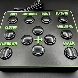 Puede incluir: Panel de control negro con letras y borde verdes, con botones etiquetados como "Putt", "Green Grid", "Scout", "Flyover", "Club Up", "Club Down", "Mulligan" y "Enter". También se ve un mando direccional y un cable USB.