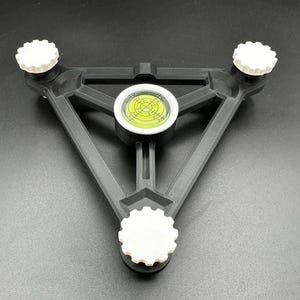 May include: A black and white levelling tool with a bubble level at the centre. The tool has three white adjustment knobs. The bubble level has a yellow liquid and a white circular frame. The tool is triangular in shape.