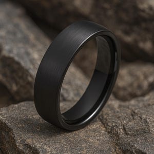 Peut inclure: Une bague noire avec une finition en métal brossé. La bague est posée sur un fond de roches grises. La bague a un intérieur lisse et arrondi et un extérieur plat et texturé.
