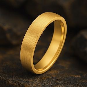 Puede incluir: Un anillo dorado con una textura cepillada. El anillo tiene un perfil plano y un interior liso. El anillo está sobre un fondo oscuro y texturizado. El anillo es de diseño simple y elegante.