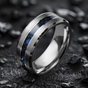 Op de afbeelding: Een zilverkleurige ring met een geborstelde metalen afwerking. De ring heeft een centrale inleg van donkerblauw en zwart gespikkeld materiaal, omlijst door twee gepolijste zilveren banden. De ring is geplaatst tegen een donkere, getextureerde achtergrond.