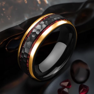 Puede incluir: Un anillo de carburo de tungsteno negro con acabado martillado, una incrustación roja y una banda dorada.