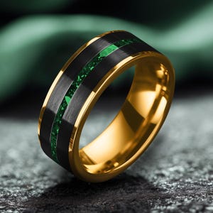 Op de afbeelding: Een goudkleurige ring met een zwarte middenband en een groene edelsteen inleg. De ring heeft een gepolijste afwerking en een modern design. De inleg bestaat uit kleine, onregelmatig gevormde groene stenen.