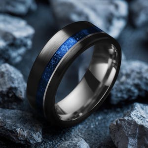 May include: A black ring with a brushed metal finish and a vibrant blue inlay. The ring is set against a backdrop of dark grey rocks, highlighting its sleek design and colour contrast. The ring's band is approximately 8mm wide.