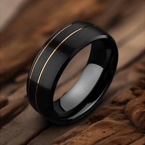 May include: A black ring with a brushed finish and two thin gold bands. The ring has a polished interior and a beveled edge. The ring is displayed on a brown, textured surface.