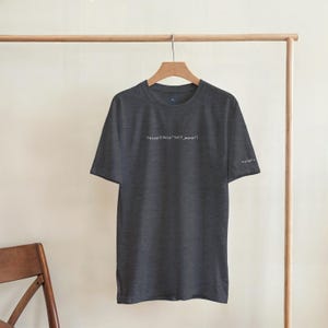 Puede incluir: Camiseta gris oscuro con el texto blanco "retrain(data:"self_worth")" impreso en el pecho. La camiseta est&aacute; colgada en una percha de madera. La manga tiene el texto "#proudly" impreso en blanco.