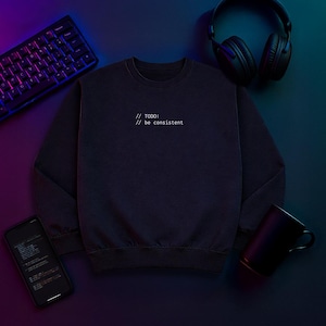 Può includere: Una felpa blu navy scuro con il testo "// TODO: // be consistent" in bianco. L'immagine include anche una tastiera, cuffie, uno smartphone e una tazza nera, il tutto su una superficie scura.