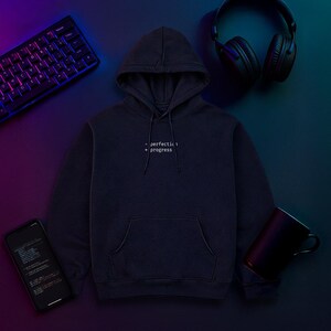 May include: A navy blue hoodie with the text "- perfection + progress" in white. The hoodie has a front pocket and a hood. A keyboard, headphones, a phone, and a mug are also visible.