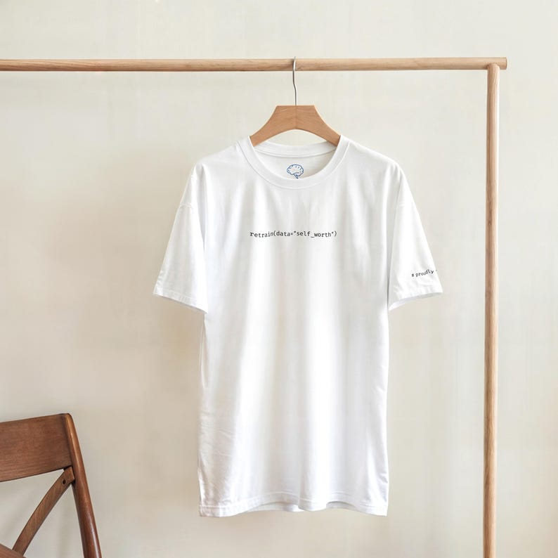 Puede incluir: Camiseta blanca con el texto "Retrain data 'self_worth'" en negro, colgada de una percha de madera. La camiseta tiene un peque&ntilde;o logotipo en la parte superior izquierda del pecho y la palabra "proudly" en la manga derecha.