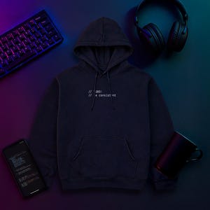 Könnte beinhalten: Ein marineblauer Kapuzenpullover mit dem weißen Text "// 000: // e consistent". Das Bild zeigt auch eine Tastatur, Kopfhörer, ein Smartphone mit Code und einen schwarzen Becher, alles auf einem dunklen Hintergrund mit lila und blauer Beleuchtung.