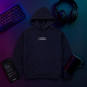 Pode incluir: Um moletom azul marinho com o texto "incremental improvement" em branco. A imagem também inclui um teclado, fones de ouvido, um smartphone exibindo código e uma caneca preta, em uma superfície escura.