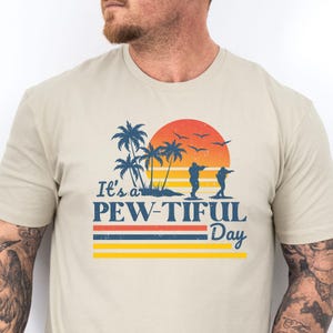 May include: Beige t-shirt with a graphic design featuring a sunset, palm trees, and two figures shooting. The text "It's a PEW-TIFUL Day" is printed below the image in blue, red, and yellow.