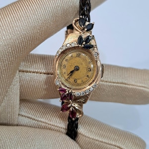 Könnte beinhalten: Vintage goldene Armbanduhr mit rundem, goldfarbenem Zifferblatt und schwarzem Lederarmband. Die Uhr hat eine dekorative Lünette mit klaren und dunklen Edelsteinen. Das Zifferblatt zeigt schwarze Ziffern und Zeiger.