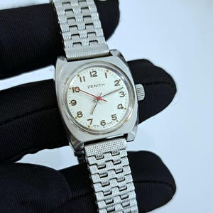 Può includere: Un orologio da polso Zenith vintage con cassa rettangolare argentata e quadrante bianco. L'orologio presenta numeri arabi neri, lancette rosse e un bracciale a maglie argentato. Lo sfondo è scuro.