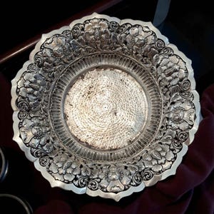 May include: Ornate silver-colored plate with a detailed floral design around the rim. The plate has a hammered texture in the center and a scalloped edge. The intricate carvings and metallic sheen suggest a decorative or serving piece.