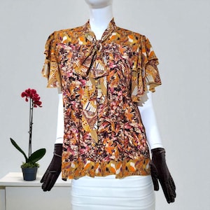 May include: A patterned blouse with a high collar and a bow tie. The short-sleeved top features a mix of orange, brown, and pink abstract designs. The blouse has ruffled sleeves and a decorative hemline. The mannequin is wearing brown gloves.