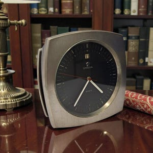 Op de afbeelding: Een vintage Omega klok met een geborsteld zilveren frame en een zwarte wijzerplaat. De klok heeft witte wijzers en markeringen, met het Omega-logo en de tekst "6 DAYS". De klok staat op 11 uur.