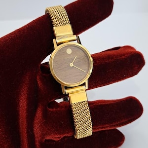 Può includere: Un orologio da polso color oro con un cinturino a maglia e un quadrante rotondo. Il quadrante dell'orologio ha un aspetto venato del legno con lancette e indici dorati. L'orologio è esposto su uno sfondo di velluto rosso scuro.