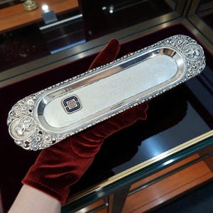 Puede incluir: Una bandeja rectangular plateada con diseños florales y figurativos ornamentados en los extremos. La bandeja tiene un borde con cuentas y un pequeño emblema con un logotipo azul y blanco. El texto es visible en la superficie de la bandeja.
