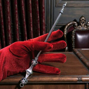 Może przedstawiać: Srebrna różdżka z ozdobnymi detalami, trzymana przez czerwoną aksamitną rękawicę. Różdżka ma palec wskazujący u góry i dekoracyjne kule. Tło obejmuje ciemną drewnianą powierzchnię i czerwone zasłony.
