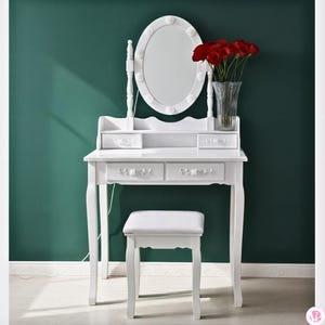 May include: A white vanity table with an oval mirror and a matching stool. The table has two drawers and a small shelf. A vase of red flowers sits on the table. The background is a dark green wall.