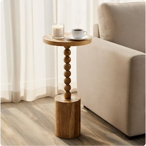 May include: A small, round wooden side table with a cylindrical base and a beaded stem. A lit candle, a cup of coffee, and a saucer sit on the tabletop. The table is next to a beige sofa and a sheer white curtain.