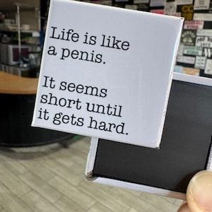 May include: A white rectangular magnet with the text "Life is like a penis. It seems short until it gets hard." The magnet is held by a hand, with a black magnetic backing.