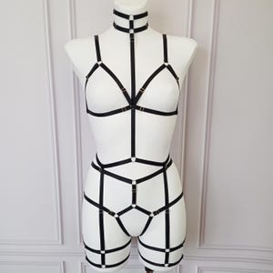 May include: Black lingerie harness set displayed on a white mannequin. The set includes a choker, bra, and shorts-style harness, all connected by straps and metal hardware. The design features geometric patterns and adjustable elements.