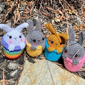 May include: Four crocheted bunny plushies in different colours. The bunnies are sitting on a bed of brown mulch. The bunnies are wearing colourful outfits.